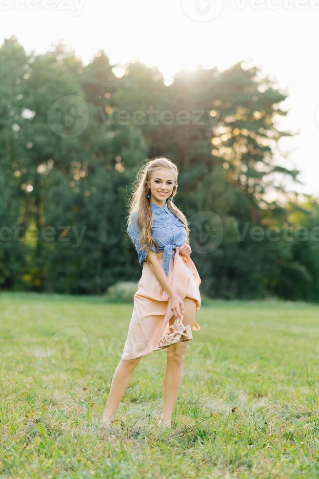 Young pretty woman in a summer dress walks through a meadow on green grass. A young girl holds shoes in her hands, showing off her beautiful leg. Summer outdoor recreation photo