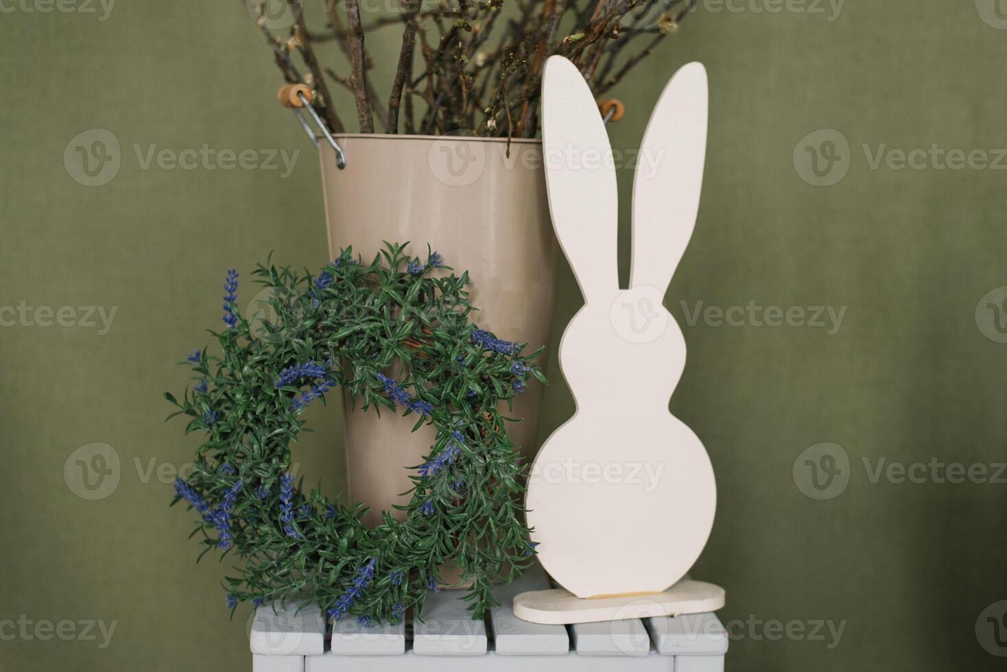 Figurine of a white rabbit stands on a wooden box next to a wreath and a bucket for Easter photo