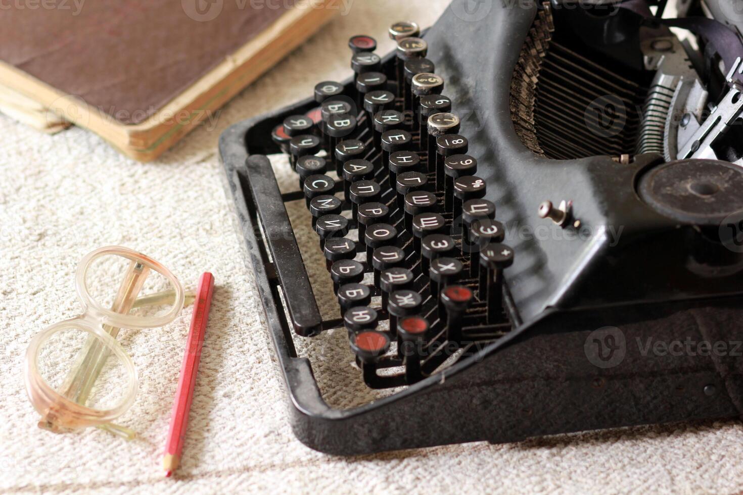 Black vintage metal type writer next to a pile of old note books, pair of eyeglasses and a red pencil on a linen tablecloth photo