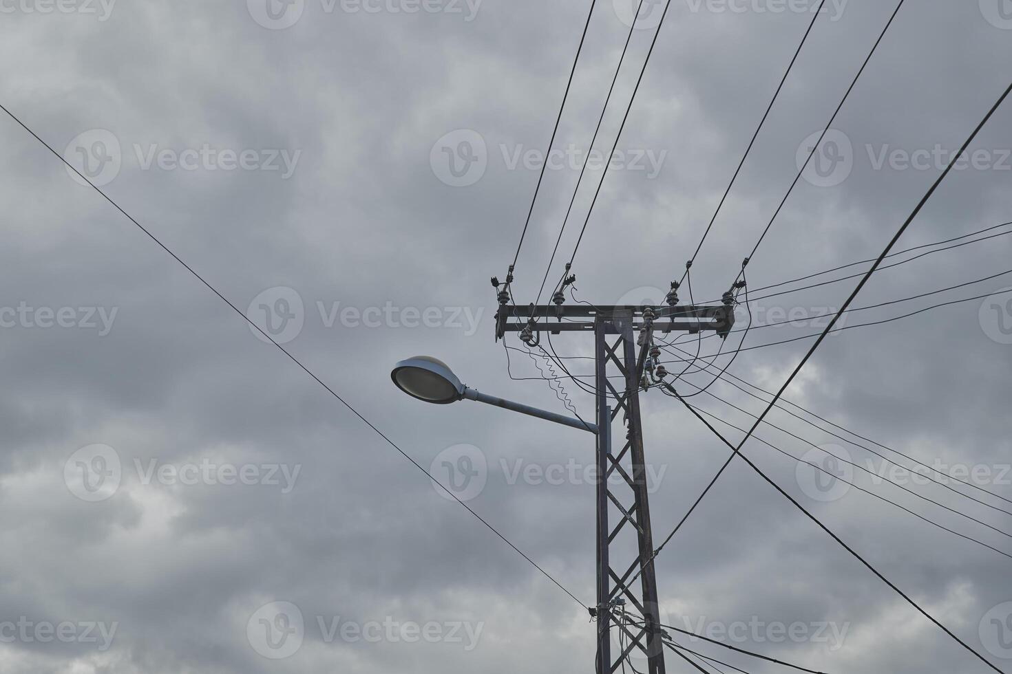 Street light mounted on a metal electrical post, accentuated by intertwining power lines against overcast sky. photo