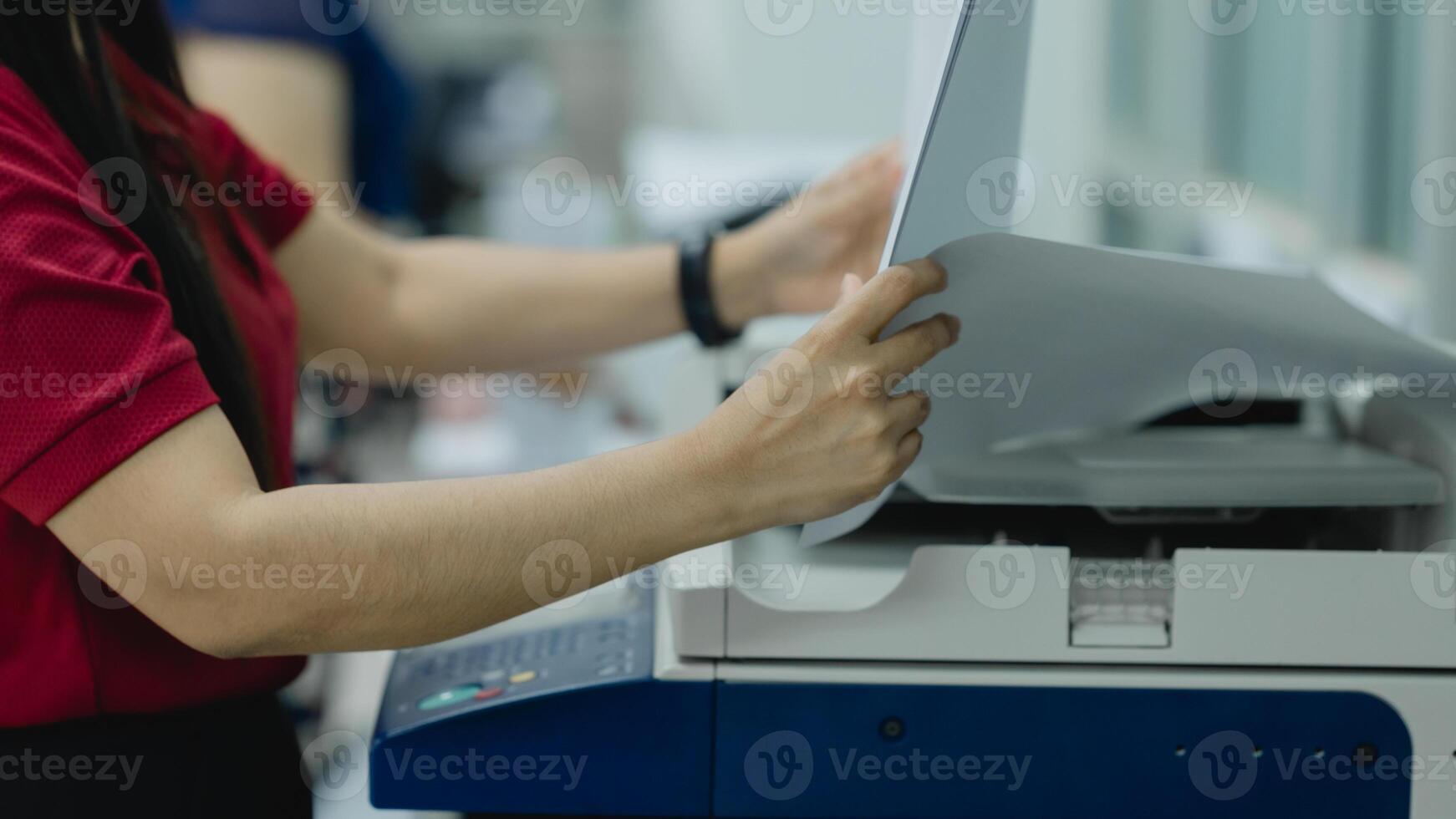 A woman is using a printer to print a document. She is wearing a red shirt and is holding a piece of paper in her hand photo