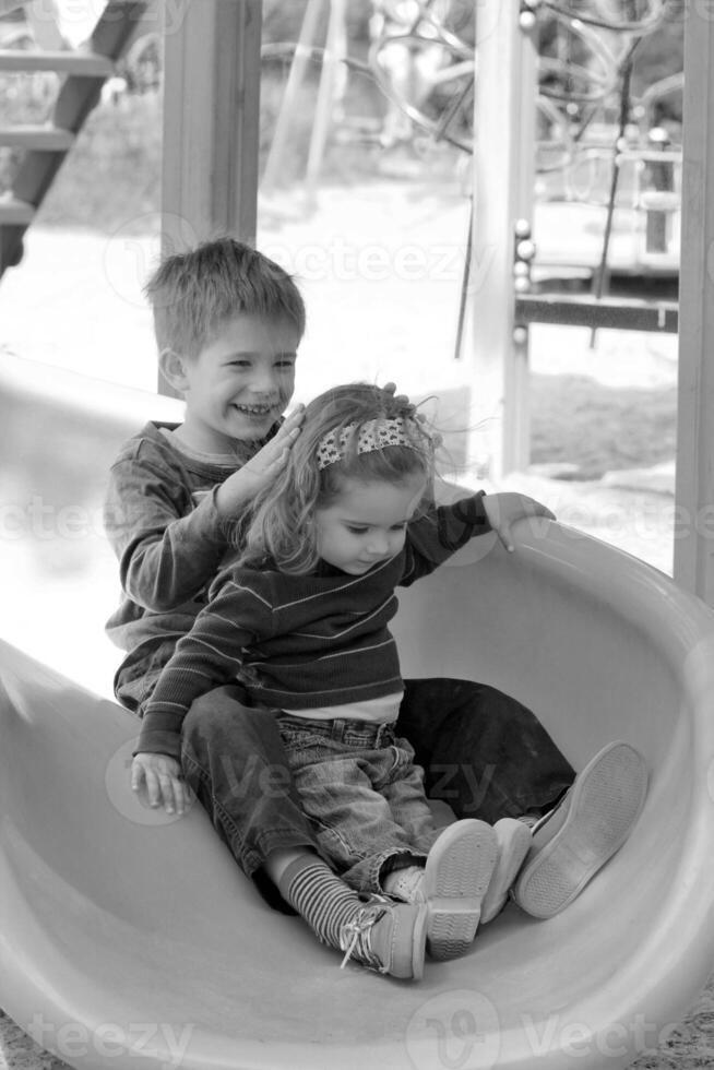 niños teniendo divertido porque de pelo levantamiento estático electricidad en el el plastico patio de recreo deslizar. negro y blanco retrato foto