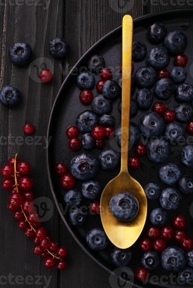 The gold metal spoon, black wood desk surface and fresh natural berries blueberries and red currant on black dish on a table. top view photo