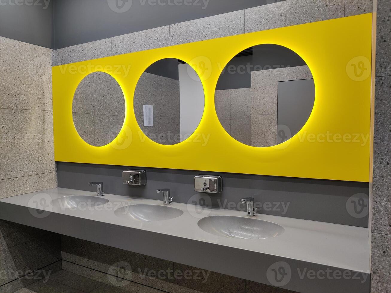 Modern bathroom interior with stone grey tiles, sink and and round yellow mirrors. bathroom with washbasin and faucet. Public bathroom in the airport or restaurant, cafe, office photo