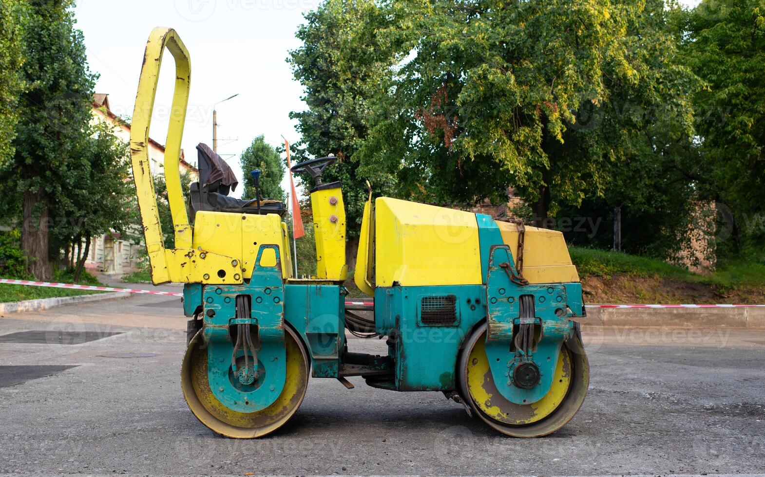 Steam roller standing idle on a path with an old stone wall and trees to the rear. road renovation, pavement patching repair photo