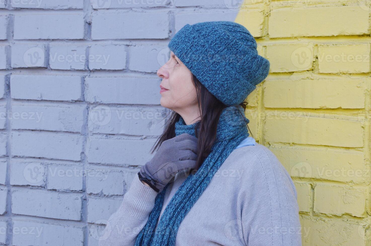 A young Caucasian brunette woman in a turquoise knitted hat and scarf stands against a brick wall and looks into the distance. photo