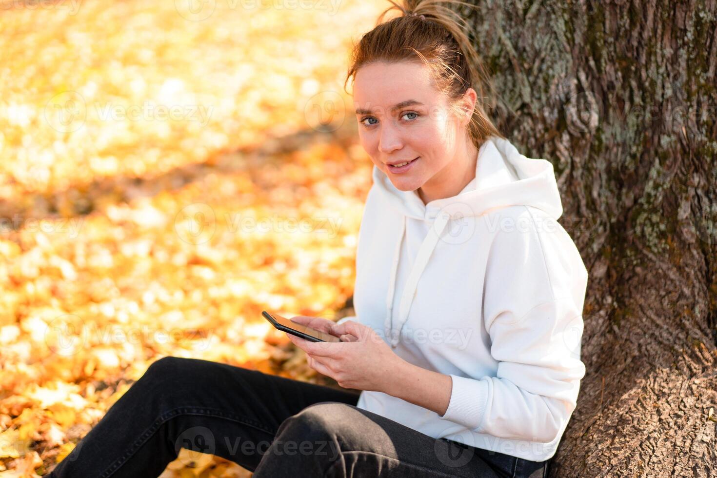 Woman in a white sweater with a hood sits on ground in the park near the tree and holding a cellphone in her hands photo