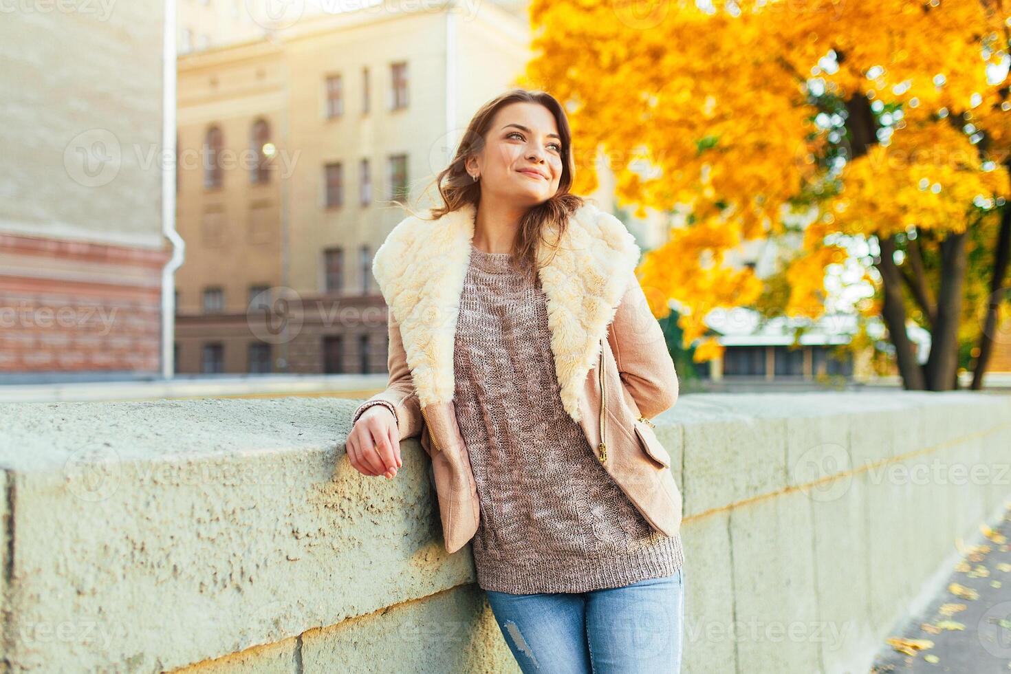 hermosa caucásico morena niña en pie calentar otoño día con antecedentes de arboles con amarillo follaje y un ciudad. foto