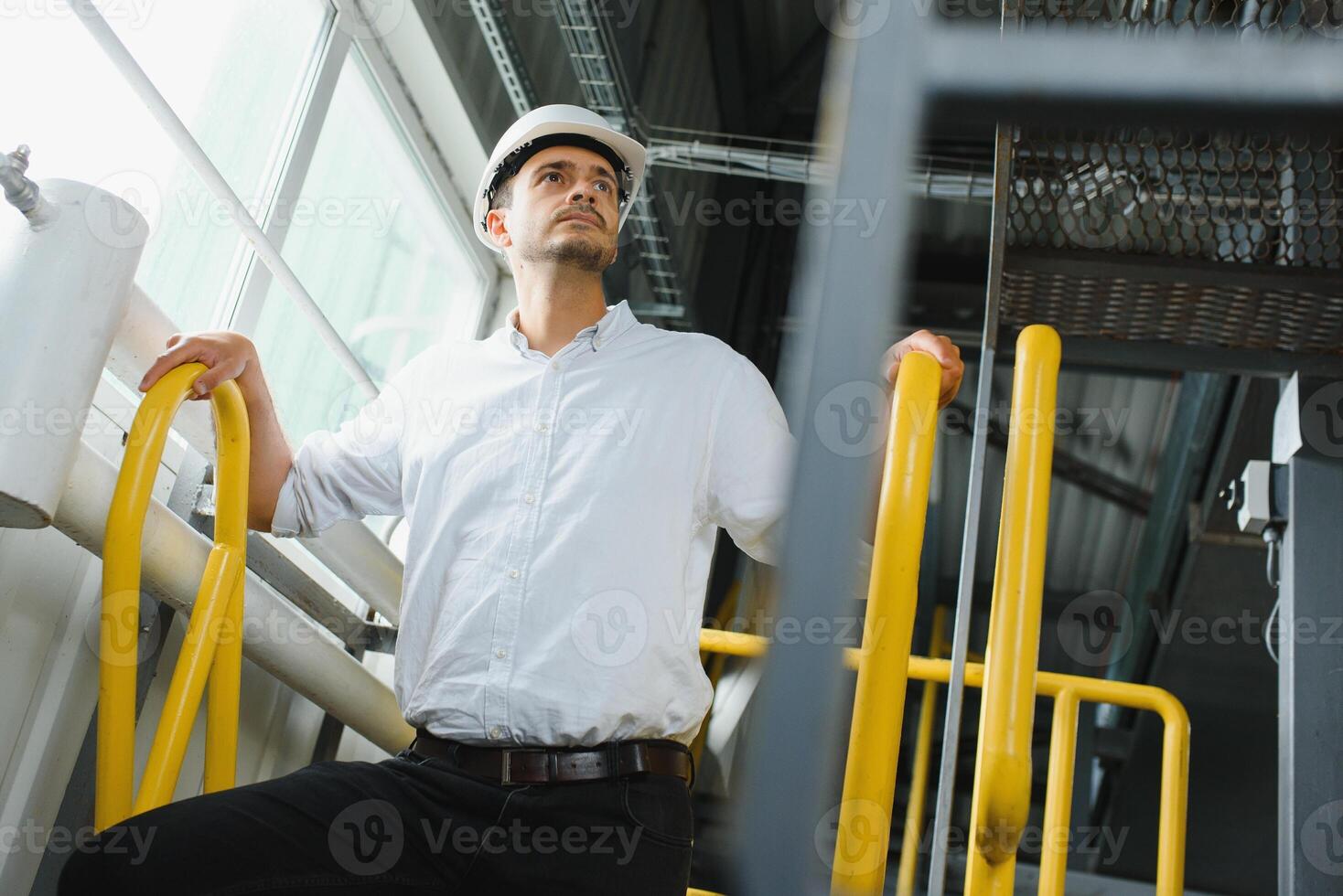 A young handsome employee of a modern factory in a white hard. In the background is a large shop for the production. photo