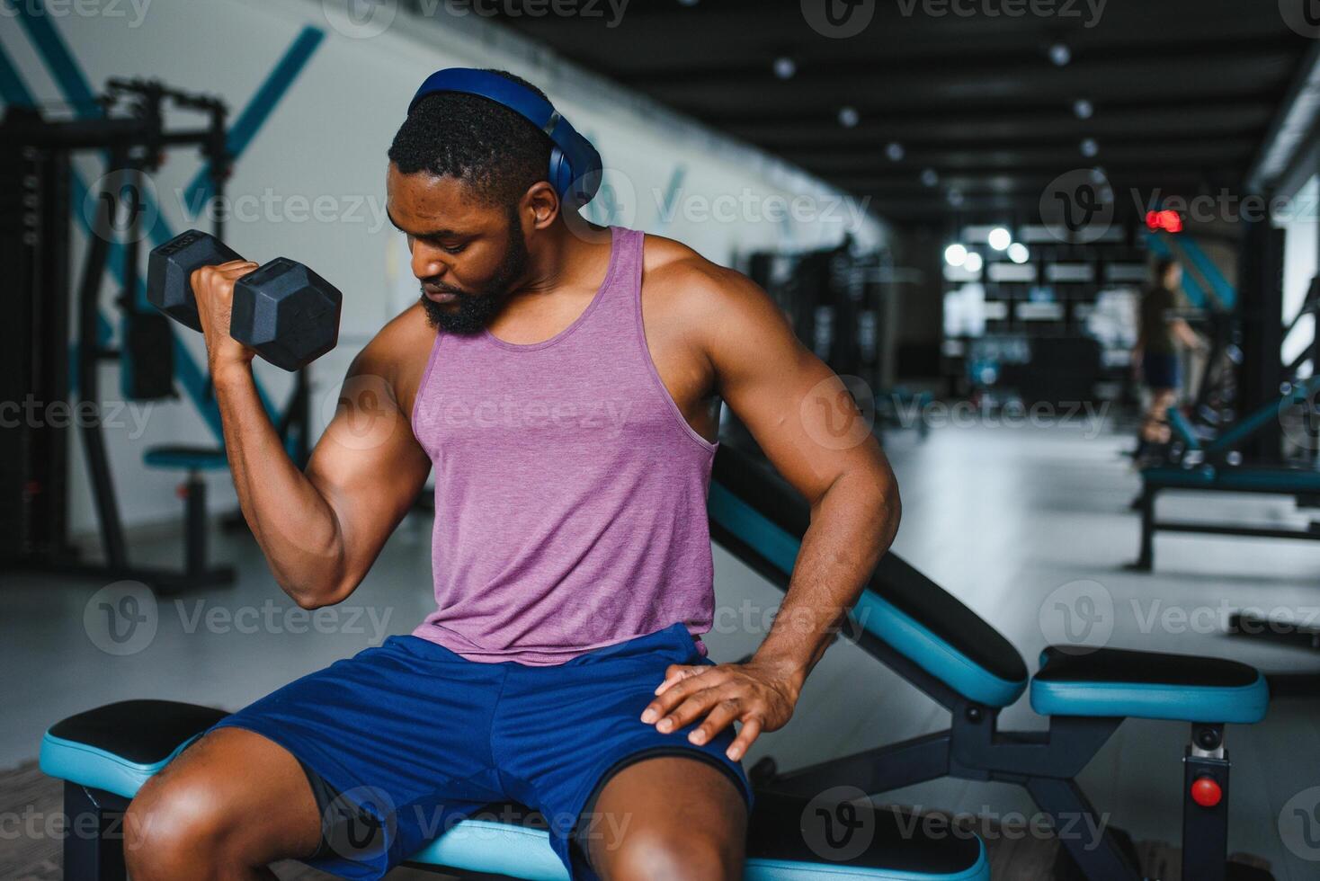 Young African American man sitting and lifting a dumbbell close to the rack at gym. Male weight training person doing a biceps curl in fitness center. photo