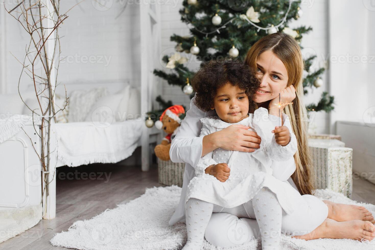 Merry Christmas and Happy Holidays Cheerful mom and her cute daughter at Christmas tree. Parent and little child having fun near Christmas tree indoors. Loving family with presents in room. photo