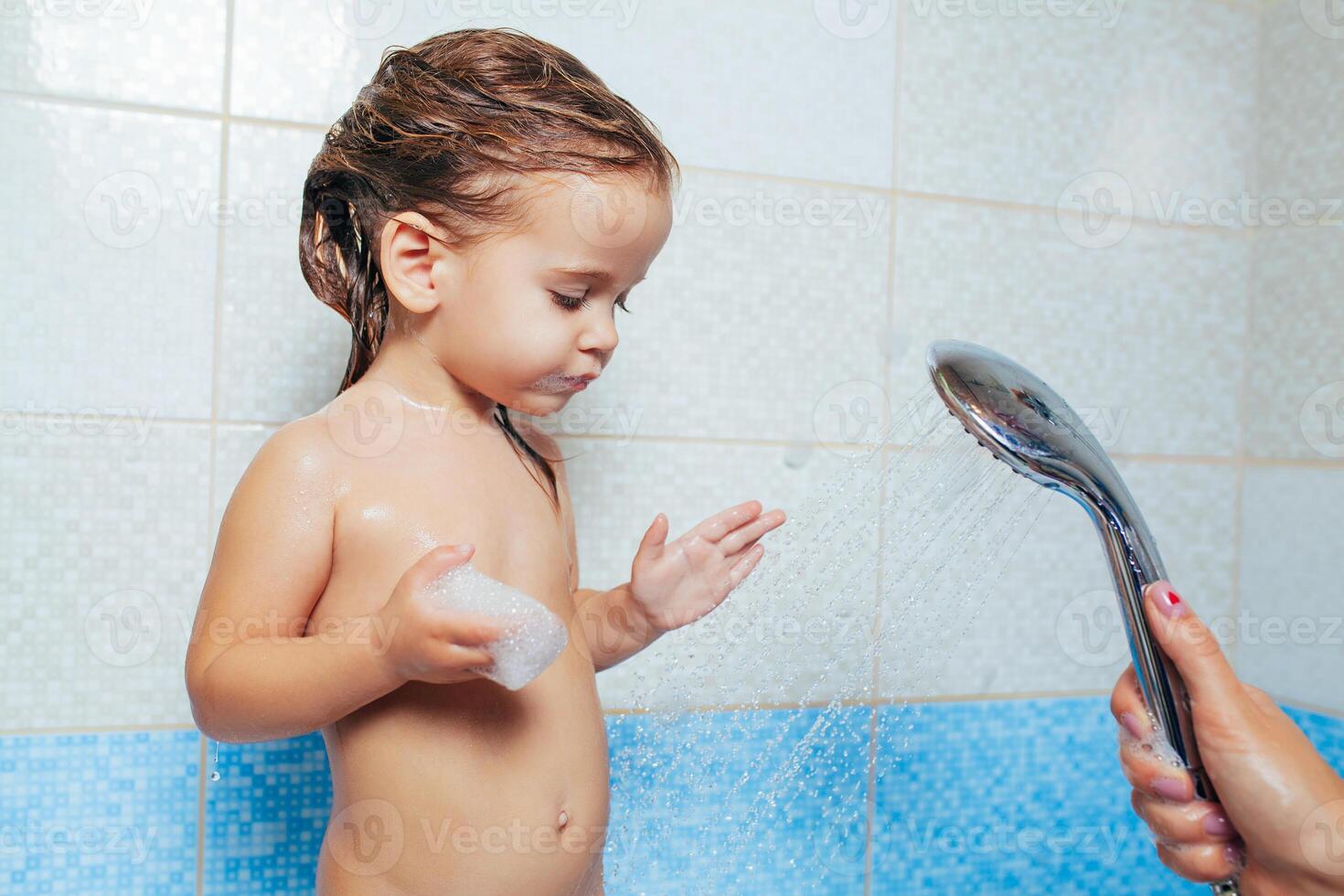 hermosa pequeño niña tomando un baño. un niño es jugado con agua y rociar desde el ducha. alegre para niños higiene. travieso hija en el baño foto