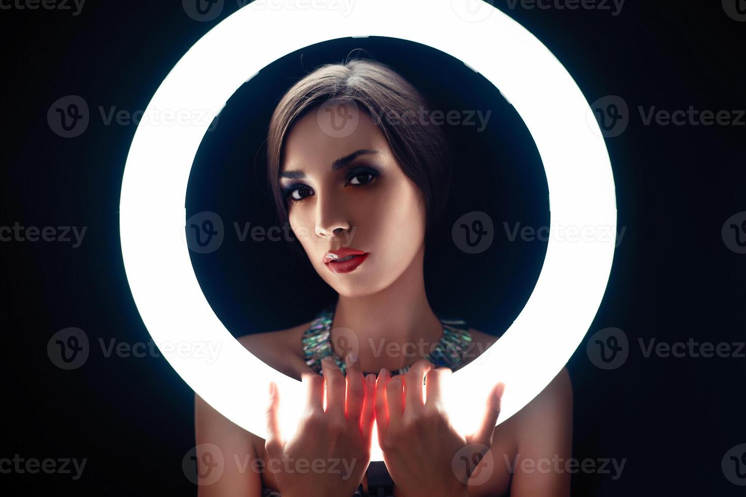 A beautiful young Caucasian girl with a beautiful make-up looks out from behind a circular lamp for portrait photo shooting