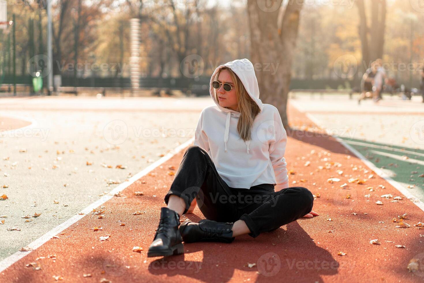 Outdoor portrait of young beautiful woman with long in sunglasses and a white hooded sweater sitting on the sportsground track photo