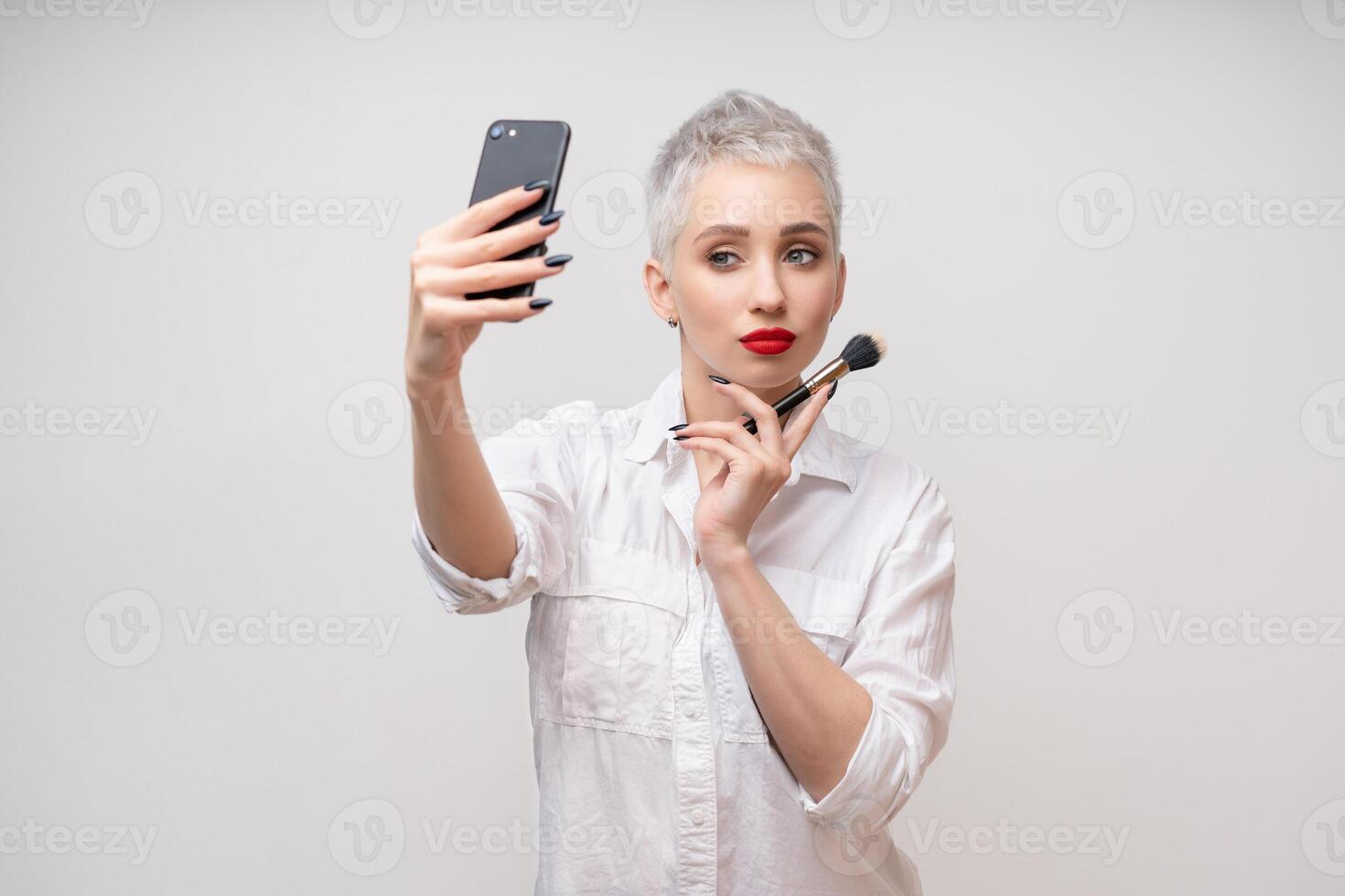 Studio portrait of beautiful trendy girl with short hair and make up holding arm on waist and taking selfie via cell phone over white isolated background photo