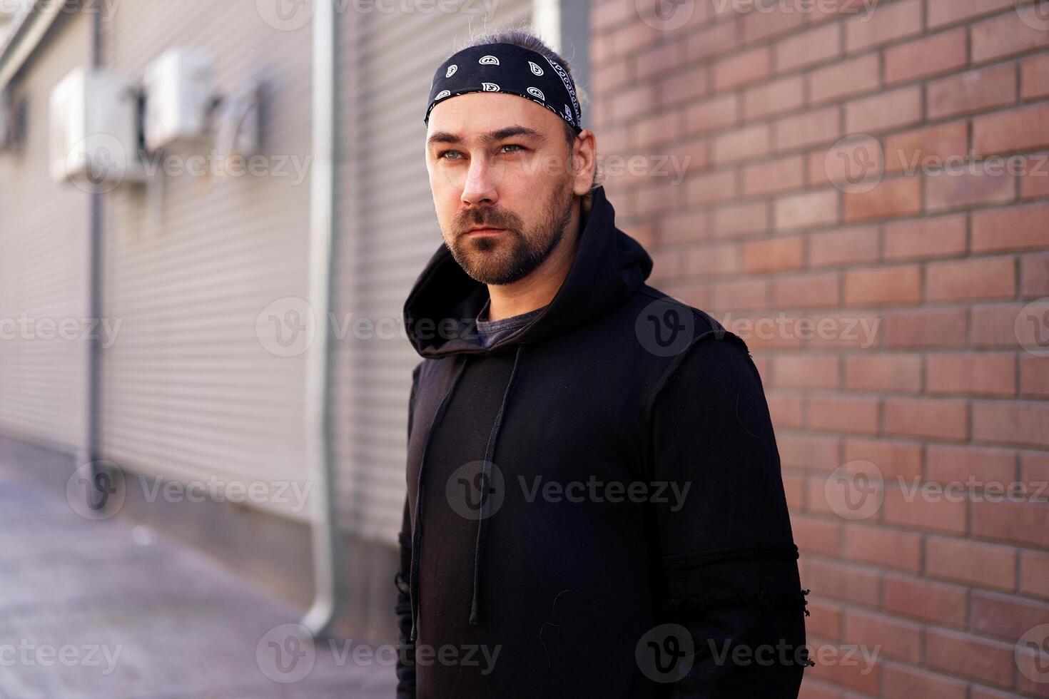 Handsome young unshaven man black hoodie and bandana stands backyard near brick walls summer day. photo