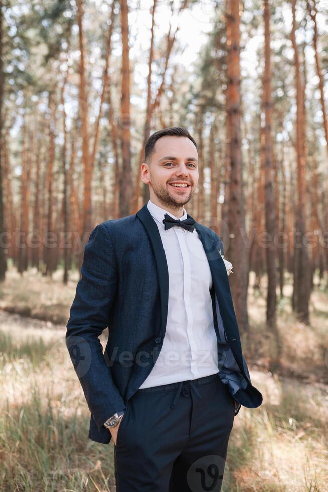 joven linda hombre en un blanco camisa negocio traje con un arco Corbata es en pie en el naturaleza en el parque y sonriente contento exitoso vida concepto foto