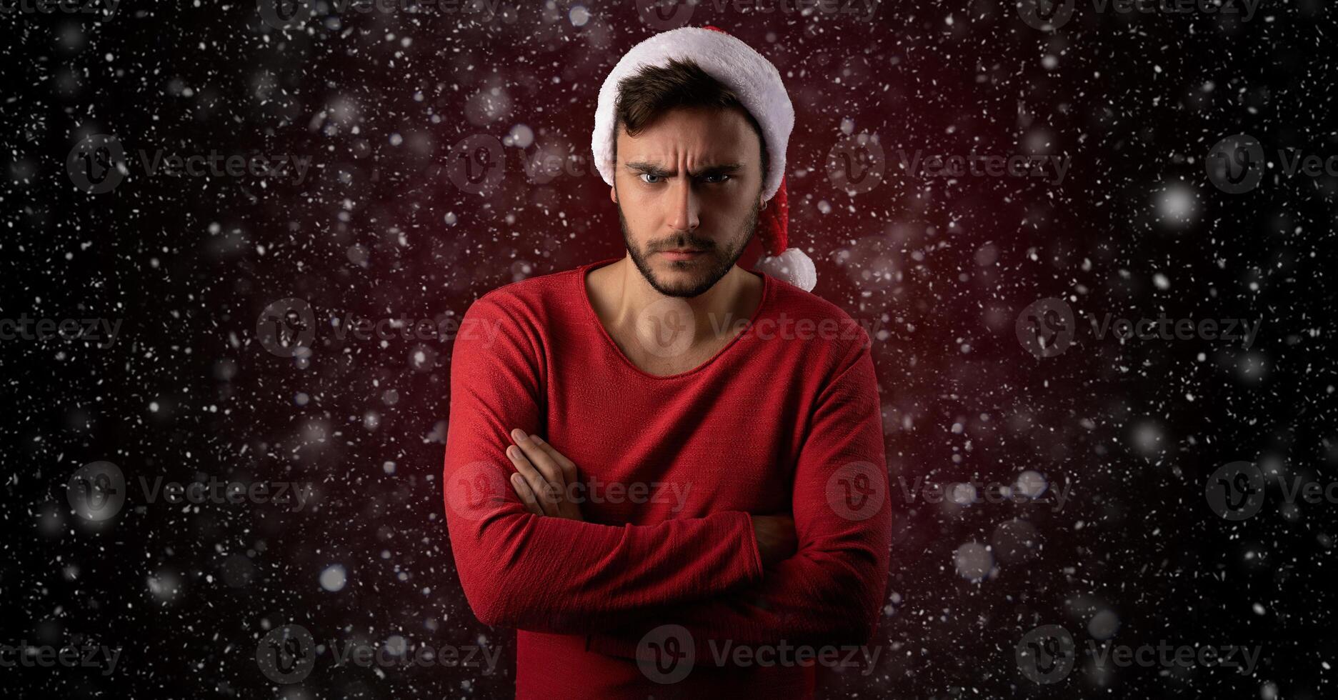 Young serious handsome caucasian guy in red sweater and Santa hats stands on red background in studio and feel angry photo