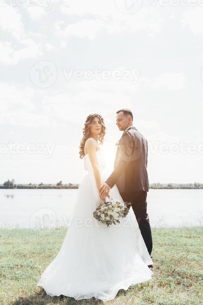 pareja vestida de boda con un ramo de flores y vegetación está en las manos contra el telón de fondo del campo al atardecer, la novia y el novio foto