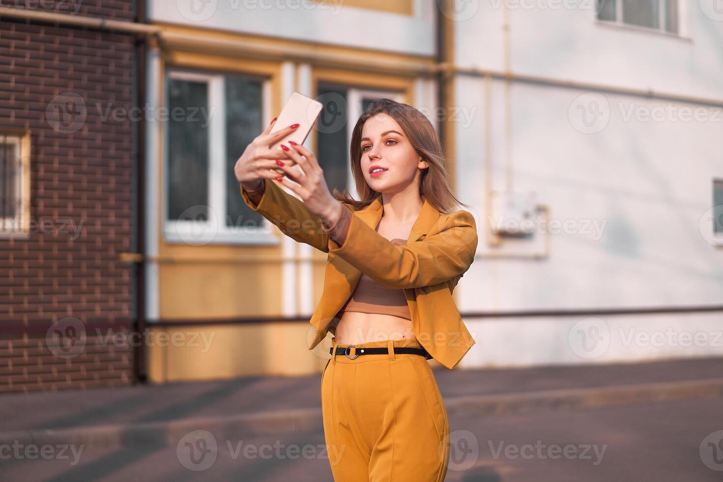 Beautiful young Caucasian girl 20 years old with white hair dressed stylish orange business suit makes selfie on the phone while standing on the street. photo