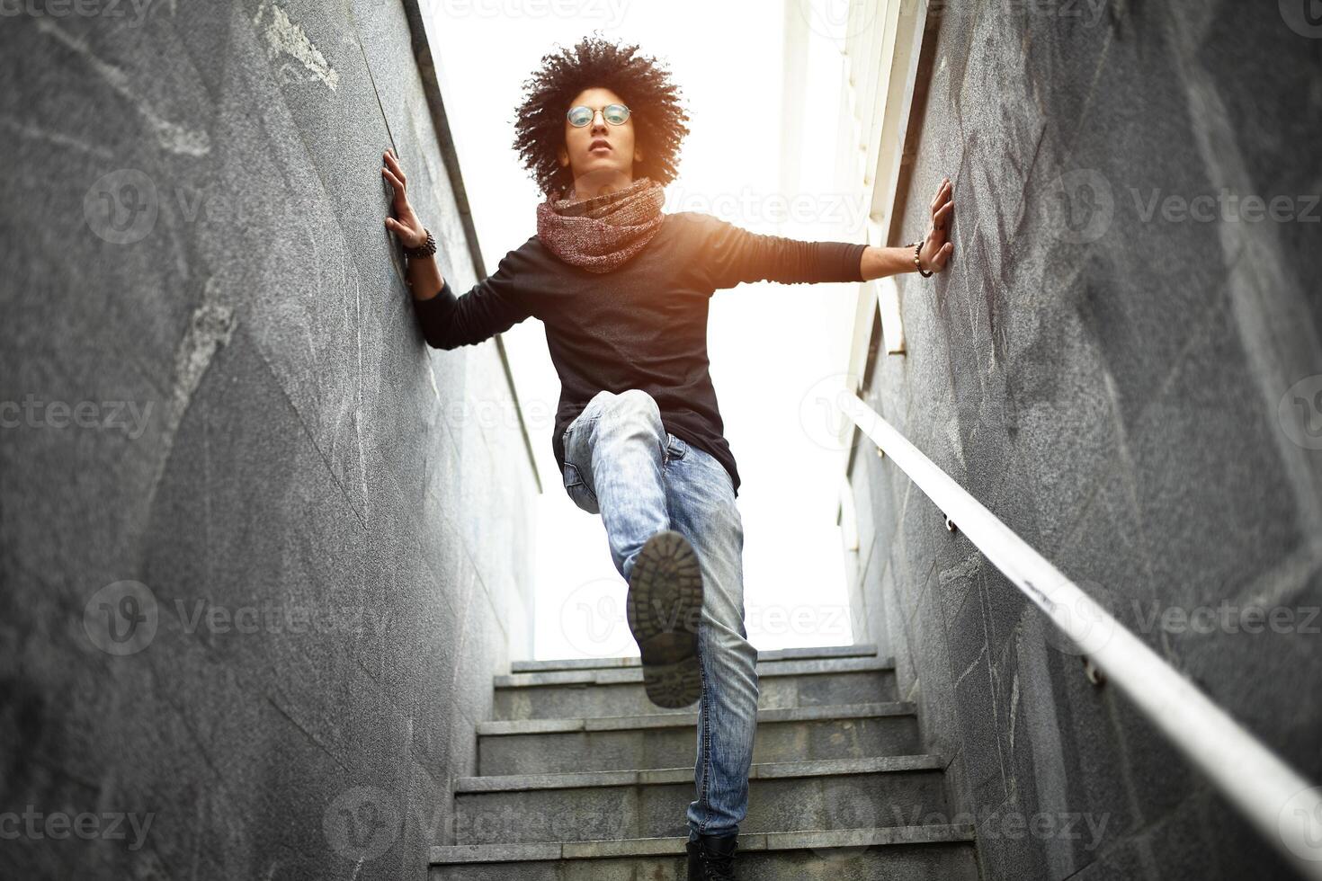 Handsome young man of mixed race with a haircut in fashionable clothes, jeans and a scarf posing against the background of a wall and stairs made of ceramic granite tiles photo