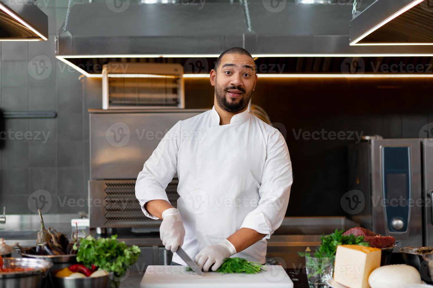 Handsome young African chef standing in professional kitchen in restaurant preparing a meal of meat and cheese vegetables. photo