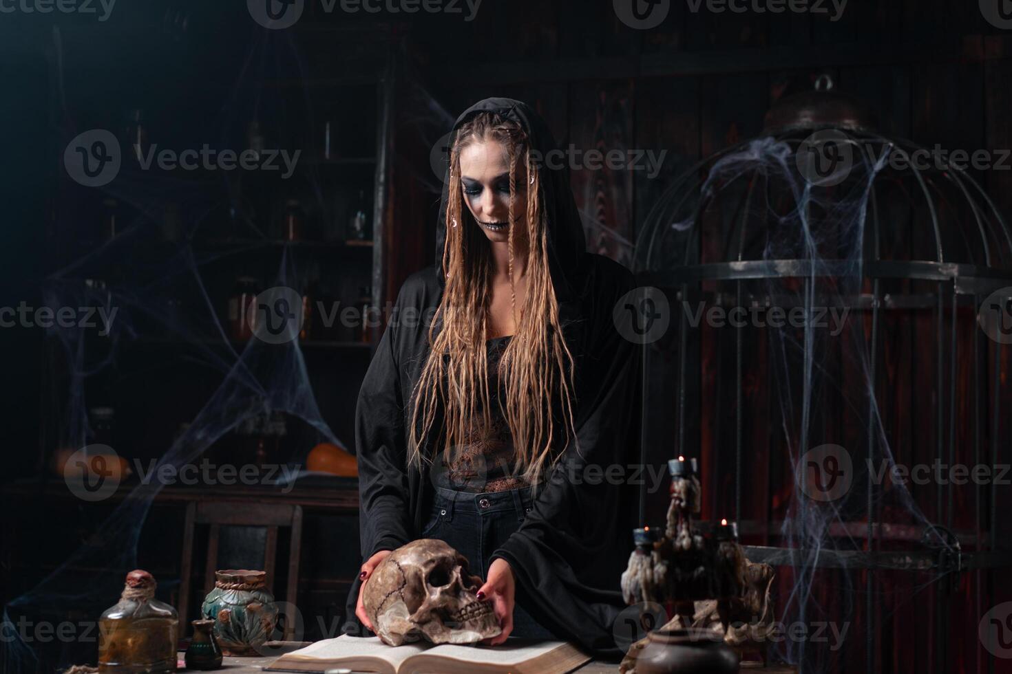 Halloween concept. Witch portrait close up with dreadlocks dressed black hood standing dark room with cage on background photo