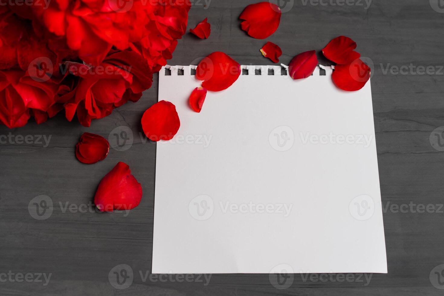 Empty sheet of paper for text next to a bouquet and red rose petals against a dark background. photo