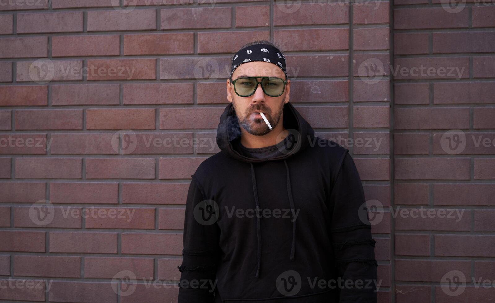 Handsome young unshaven man black hoodie and bandana stands backyard near brick walls summer day. photo