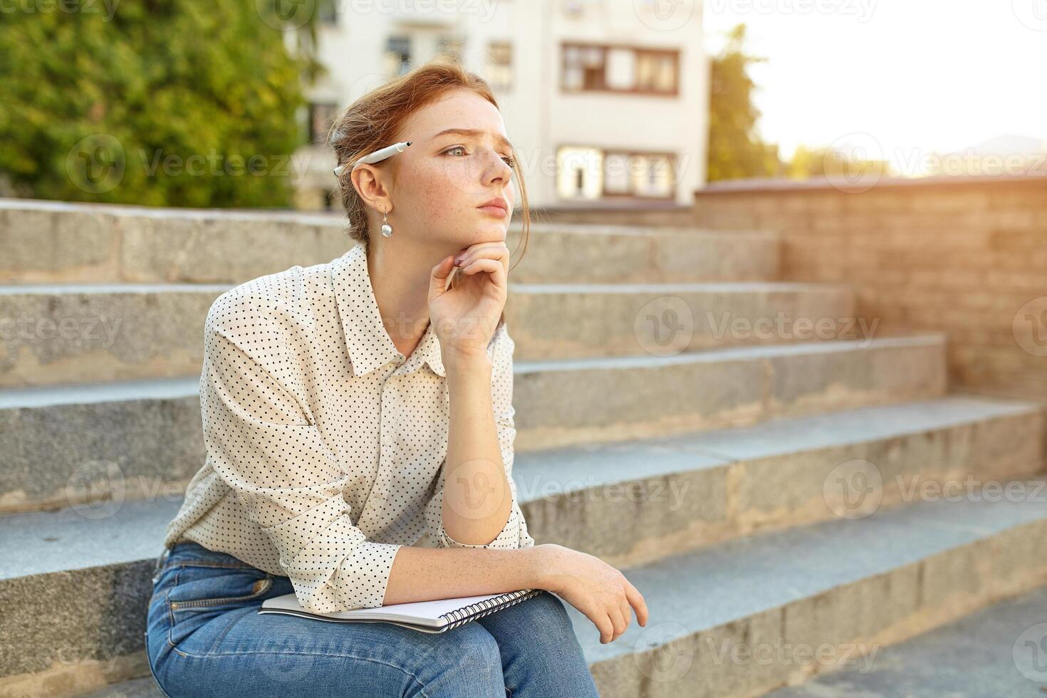 young beautiful student writes an essay in her notebook sitting on the steps stairs outdoor Red-haired girl with freckles photo