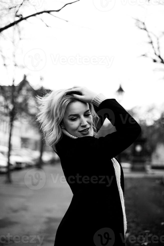 Blonde Woman in Black Coat and Scarf Standing in Front of Building. Elegant Urban Portrait of Stylish Lady. City Fashion and Architecture Concept. Chic Female Model Outdoors photo