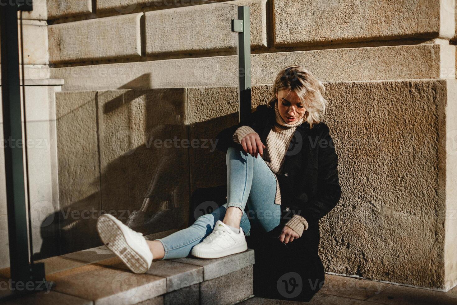 Stylish Woman in Black Coat and White Shoes Sitting on Stone Ledge. Fashionable Urban Lifestyle Portrait of Young Female. Casual Street Style Concept with Woman Relaxing Outdoors photo