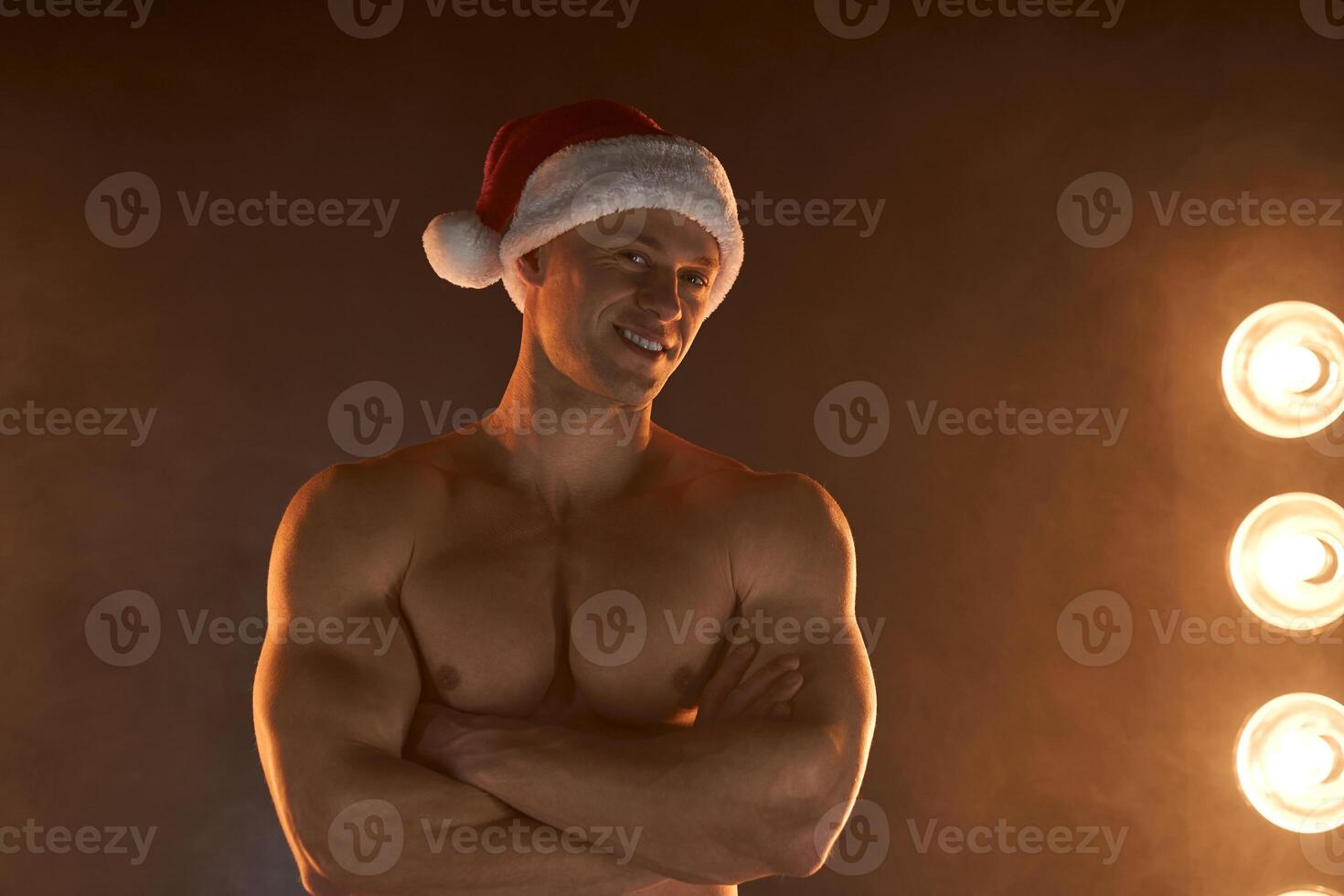 Portrait of muscular man wearing Christmas Santa hat, folded hands on smoky background photo