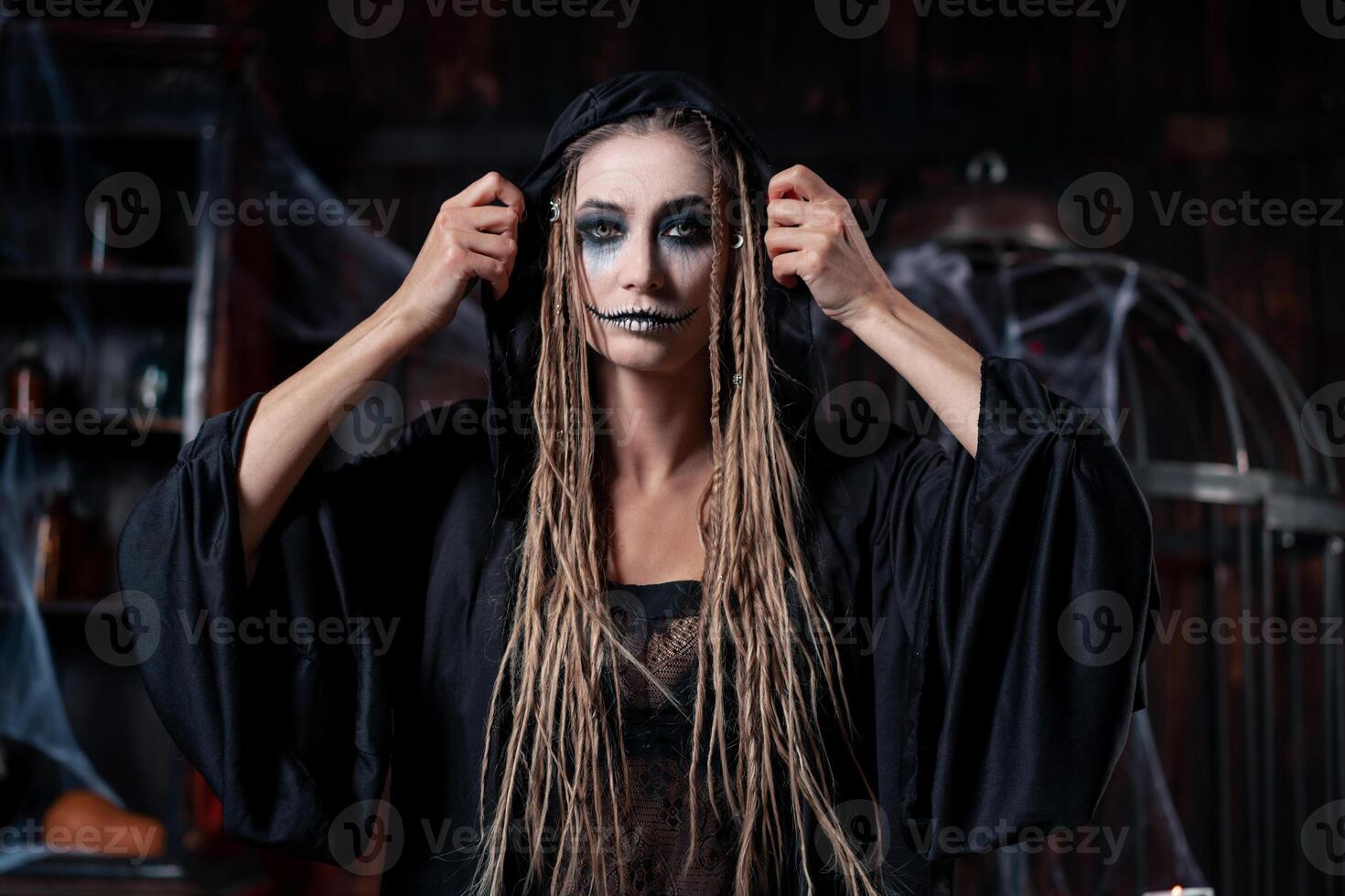 Halloween concept. Witch portrait close up with dreadlocks looking camera dressed black hood standing dark room with cage on background photo