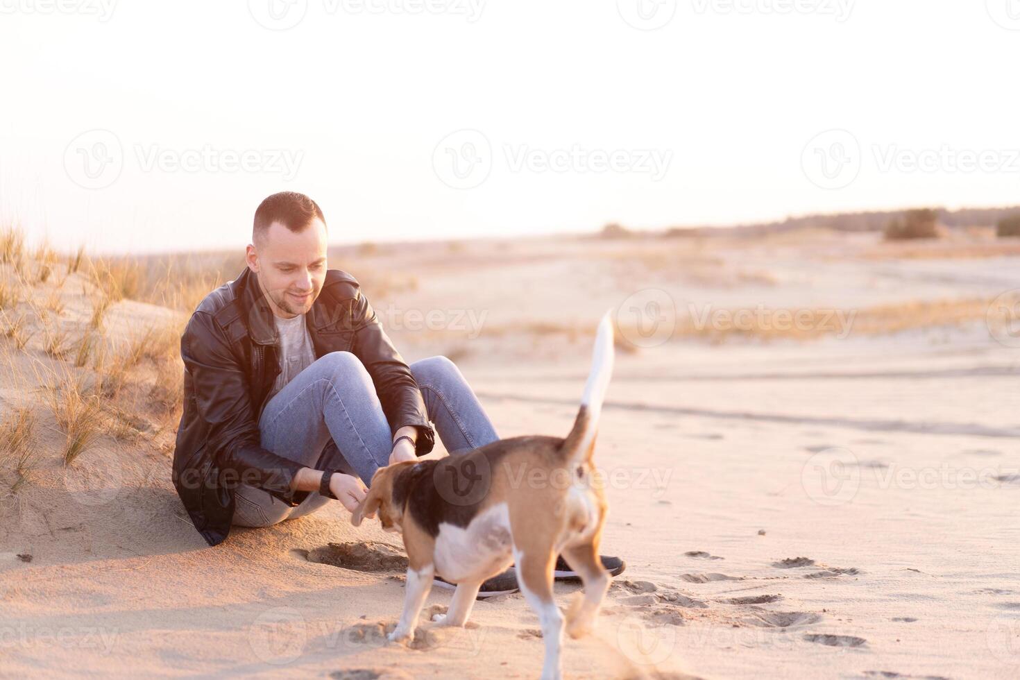 un joven caucásico hombre vestido negro cuero chaqueta y azul pantalones se sienta en arenoso playa siguiente a su amigo el perro beagle criar. foto