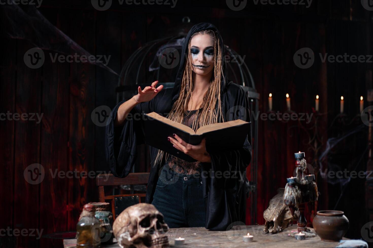 Halloween concept. Witch dressed black hood with dreadlocks standing dark dungeon room use magic book for conjuring magic spell photo