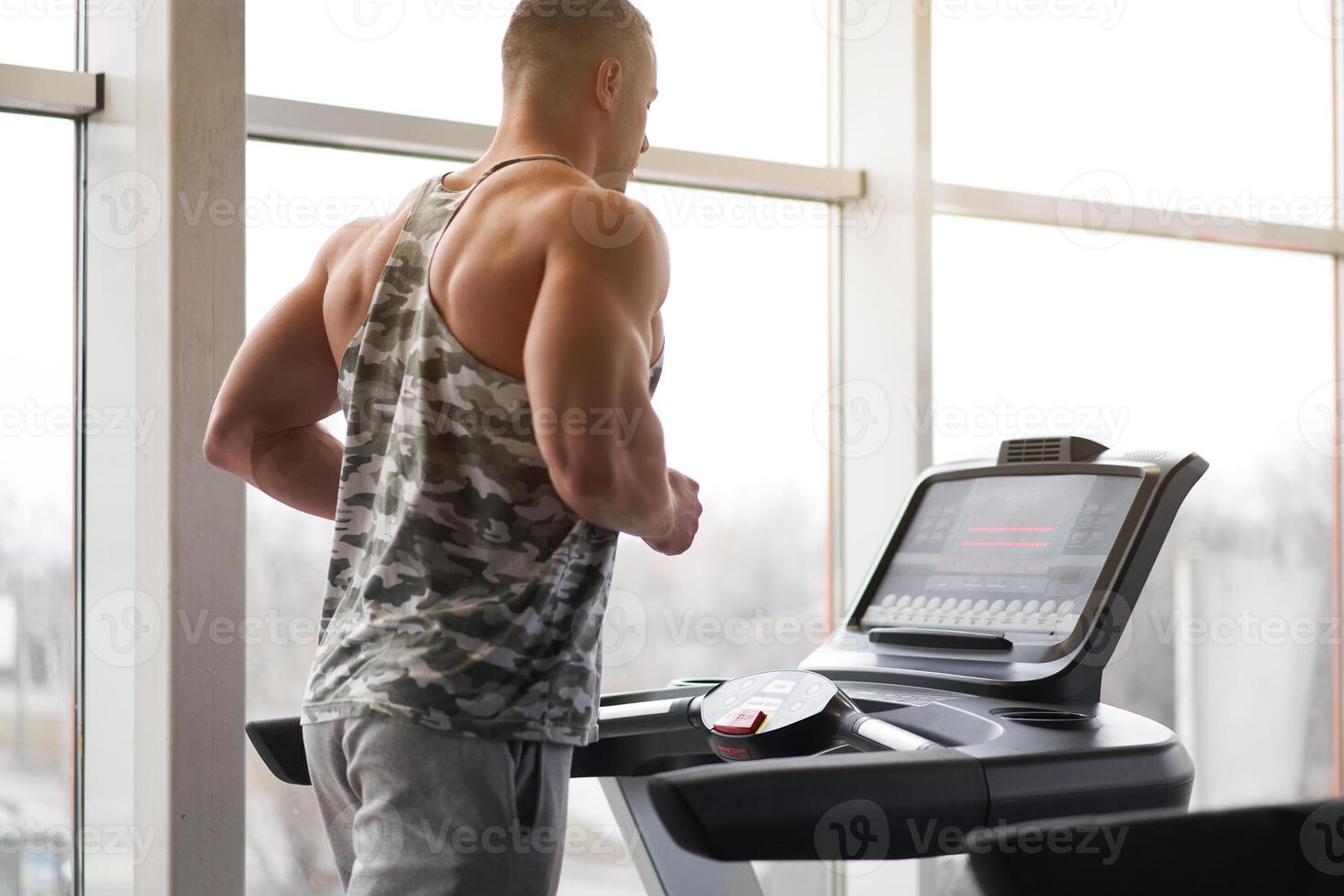 Muscular athletic bodybuilder fitness model running treadmill gym near big window photo