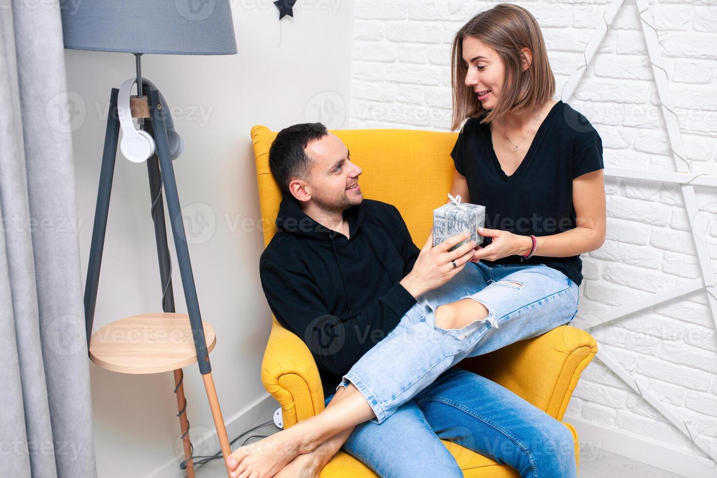Portrait of couple young in a cozy room. A man sits on a yellow chair. A woman sits on the lap of a man and holds a gift photo