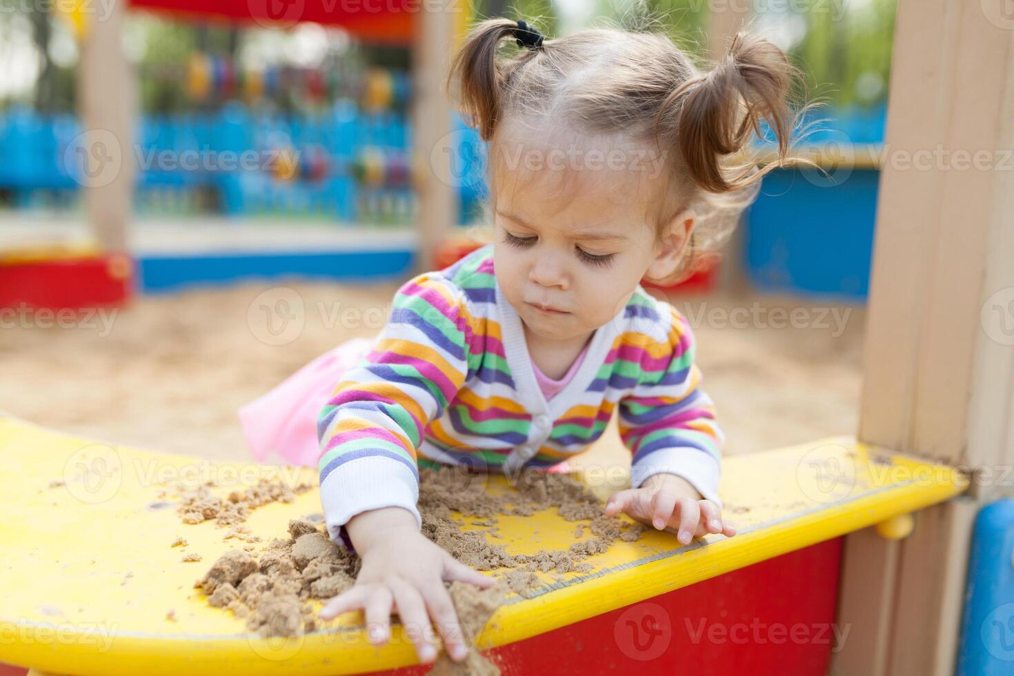 a little girl with two tails is dressed in a striped colorful jacket is playing in the sandbox on the playground photo