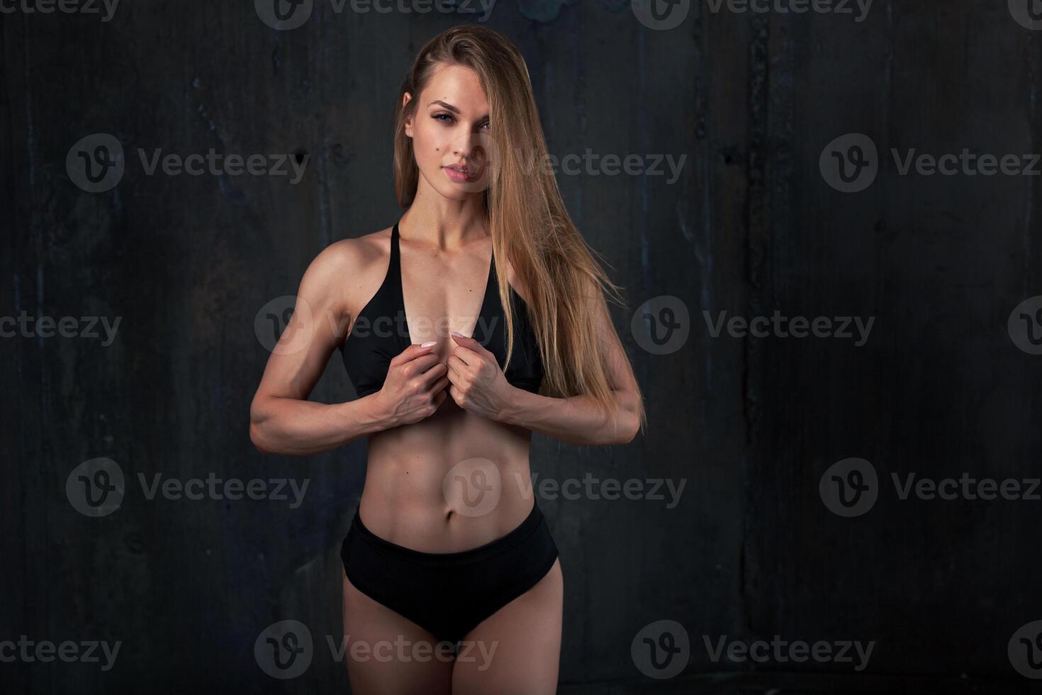 Image of muscular young female athlete wearing black sport wear standing looking in camera on dark background. Woman bodybuilder resting after workout. photo