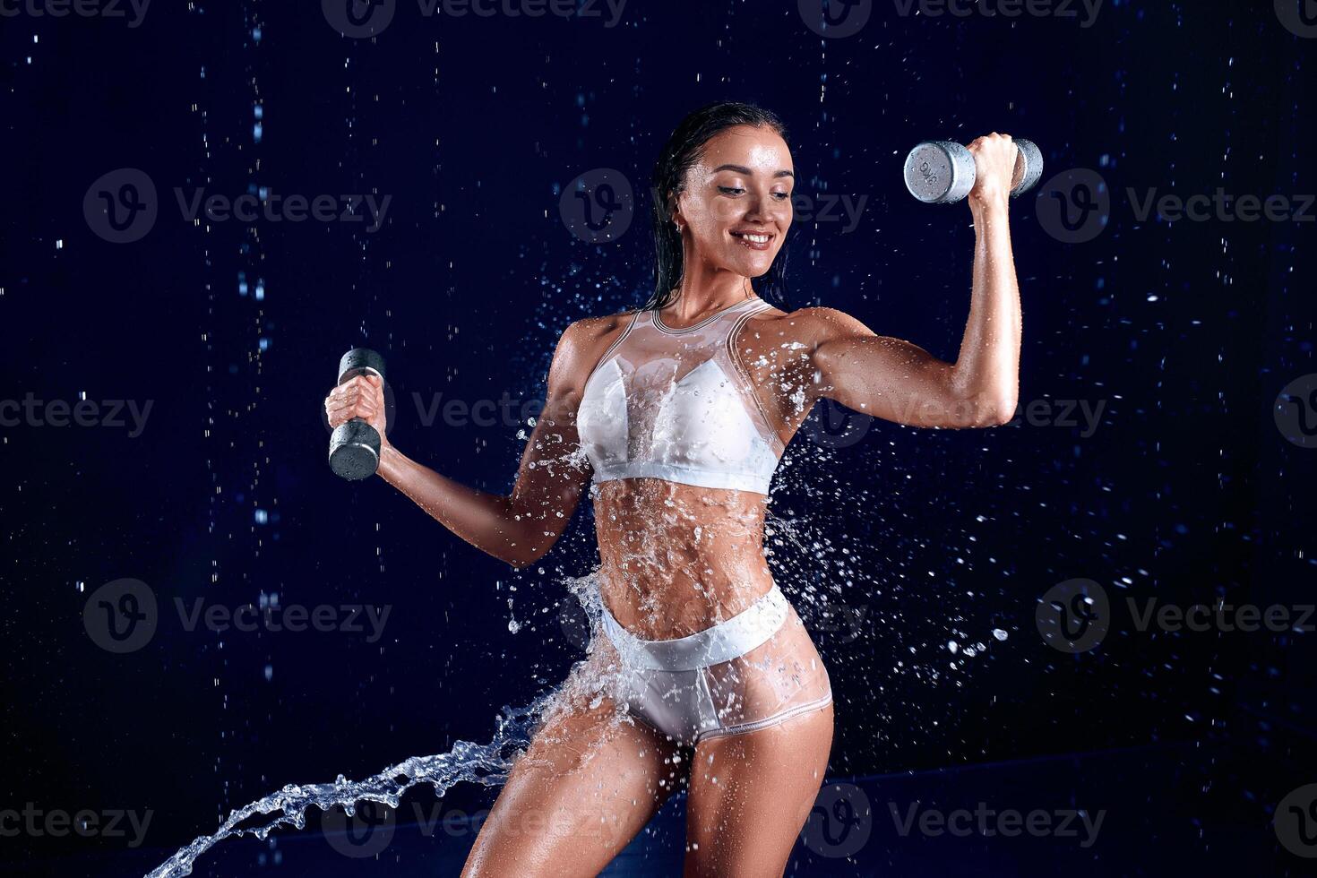 hermosa joven niña en ropa de deporte poses con pesas en agua estudio. gotas de agua untado acerca de su aptitud cuerpo. el Perfecto figura en el antecedentes de agua salpicaduras foto