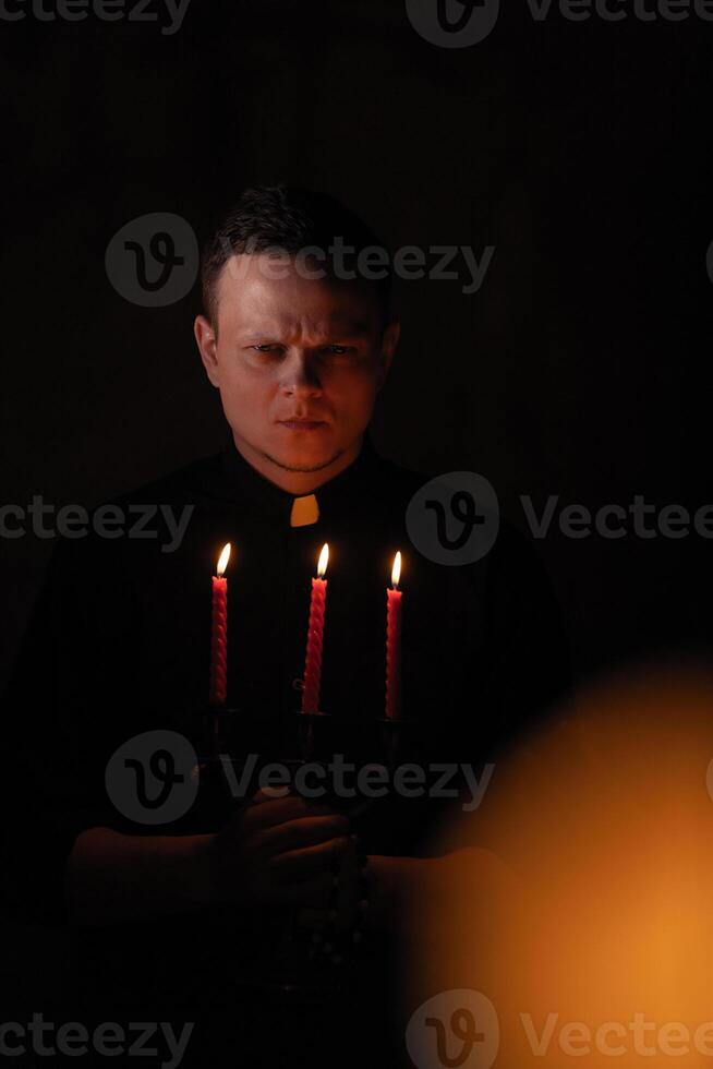 retrato de hermoso católico sacerdote o pastor con perro cuello, oscuro rojo antecedentes. con Tres rojo vela en el mano foto