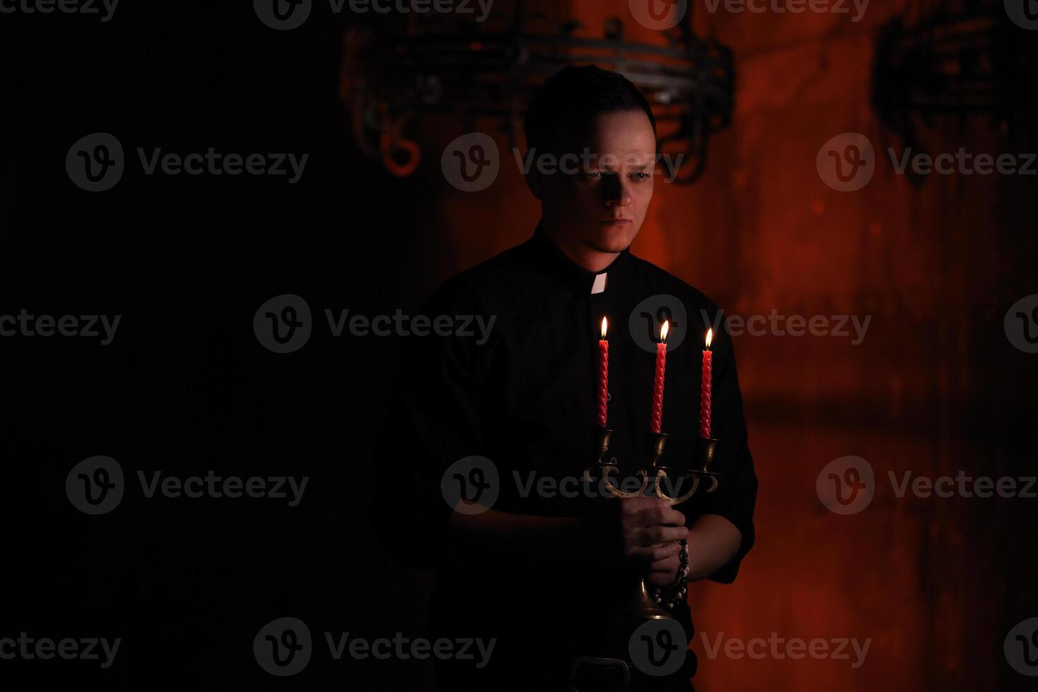 Portrait of handsome catholic priest or pastor with dog collar, dark red background. with three red candle in the hand photo