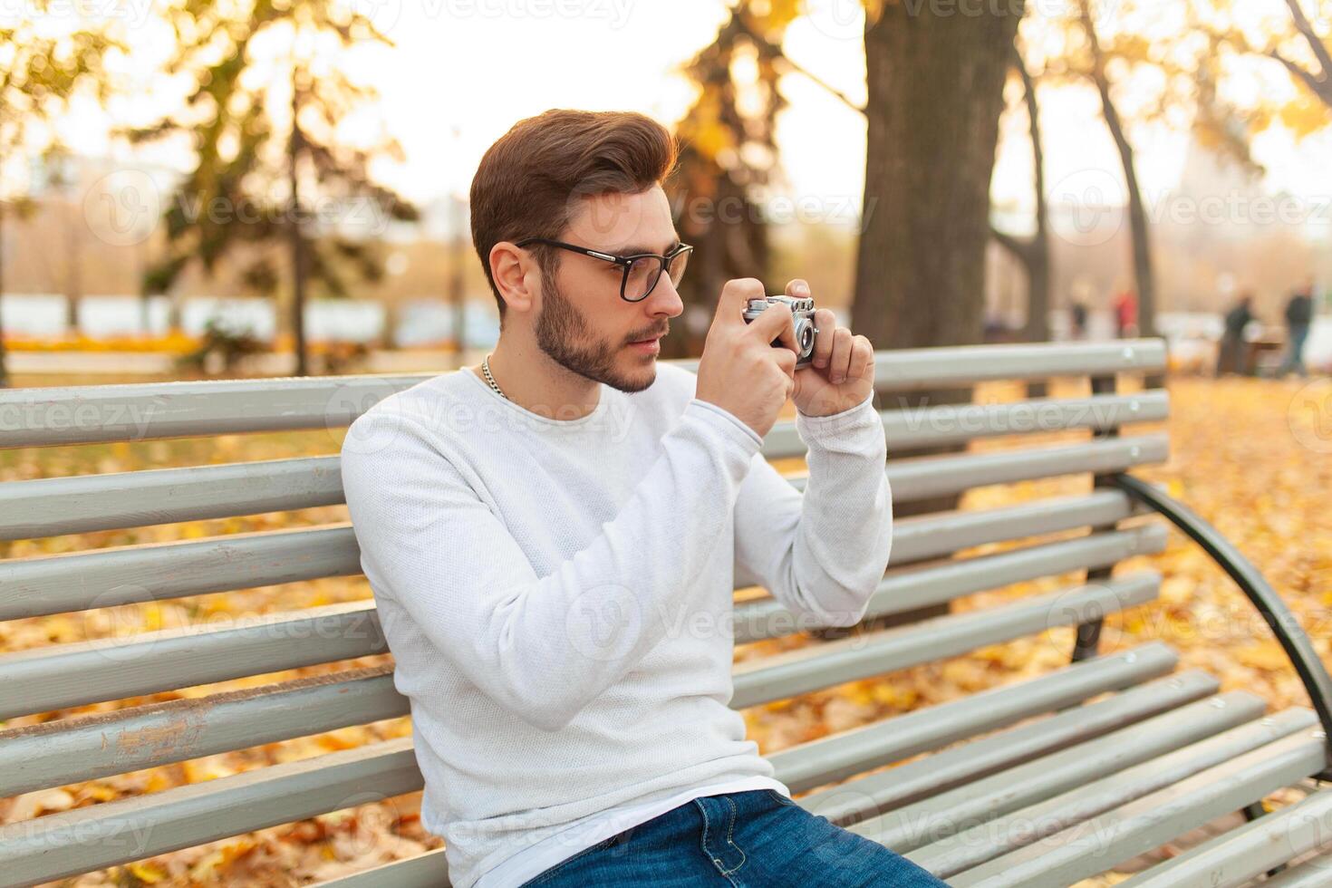 Young handsome hipster guy walks in a beautiful autumn park on the background of yellow leaves in warm sunny weather and takes pictures on a pig film camera. Autumn leisure time. Creative youth photo