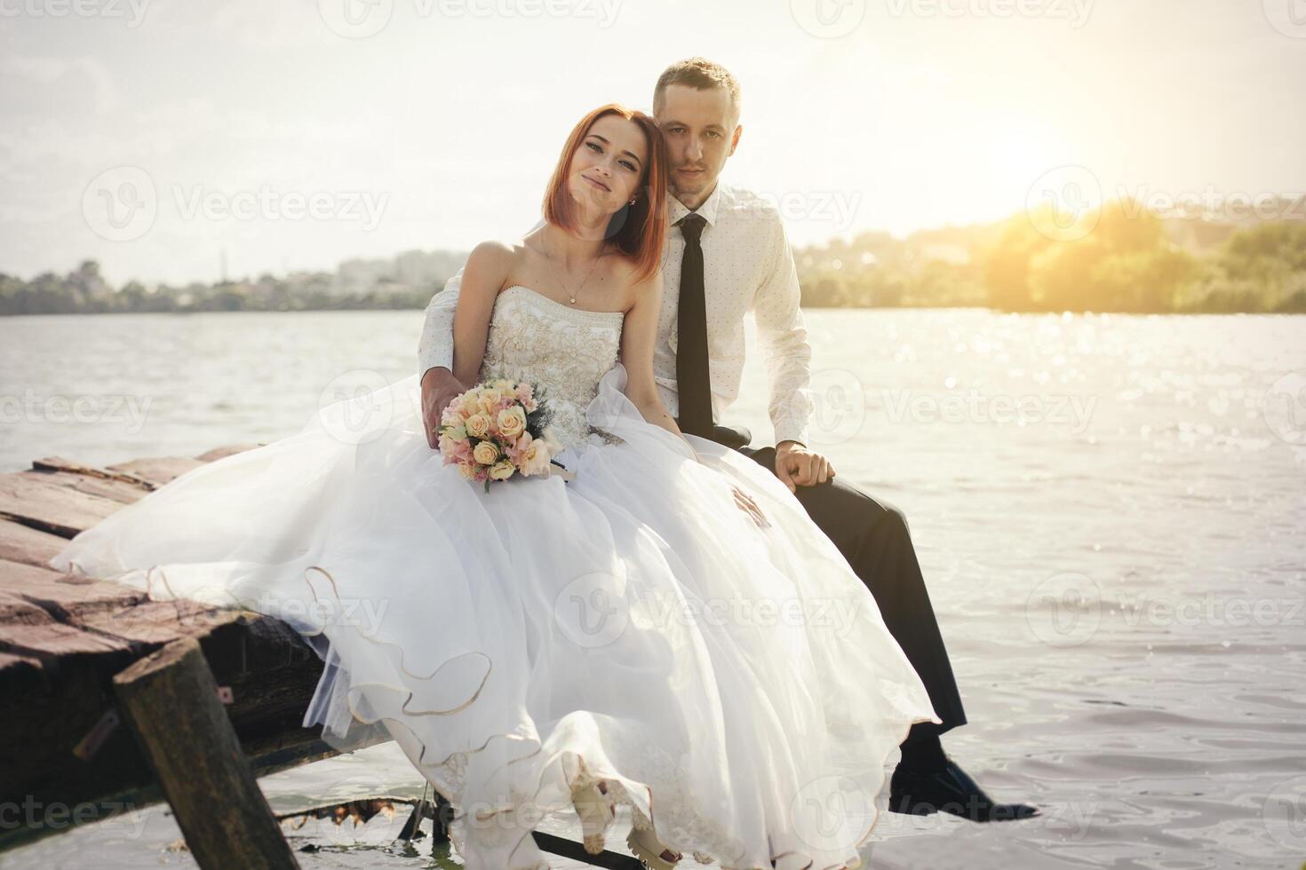 Boda Pareja sentado en puente cerca lago en puesta de sol a Boda día. novia y novio en amor foto