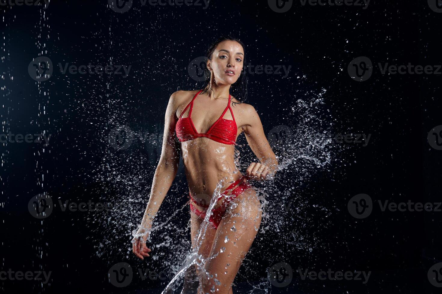 hermosa joven niña en rojo ropa de deporte en agua estudio. gotas de agua untado acerca de su aptitud cuerpo. el Perfecto figura en el antecedentes de agua salpicaduras foto