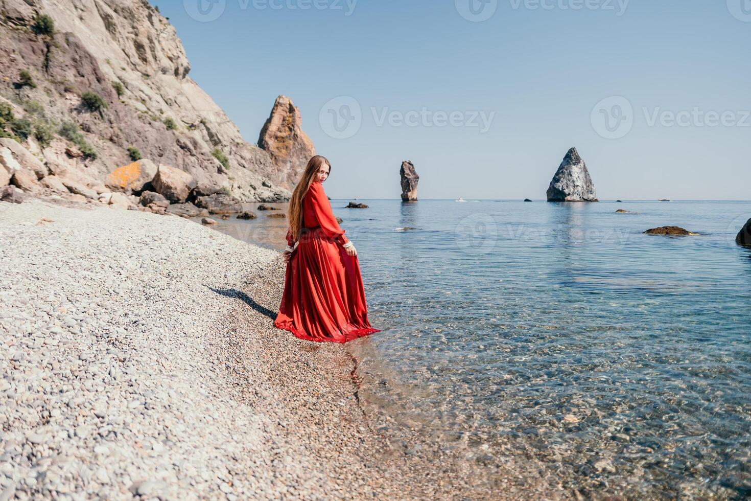 Woman travel sea. Happy tourist in red dress enjoy taking picture outdoors for memories. Woman traveler posing on the rock at sea bay surrounded by volcanic mountains, sharing travel adventure journey photo