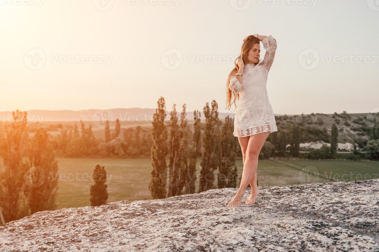 Happy woman in white boho dress on sunset in mountains. Romantic woman with long hair standing with her back on the sunset in nature in summer with open hands. Silhouette. Nature. Sunset. photo