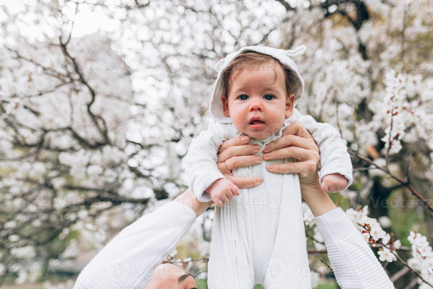 retrato de contento alegre niño en blanco ropa terminado árbol flores florecer antecedentes. familia jugando juntos afuera. mamá alegremente sostener pequeño hija foto