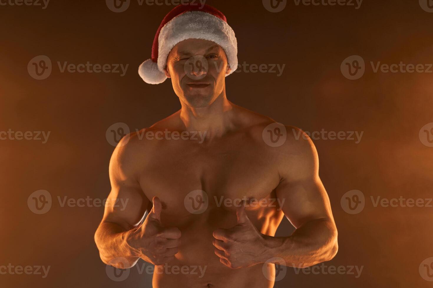 Portrait of muscular man wearing Christmas Santa hat, showing thumb up on smoky background photo